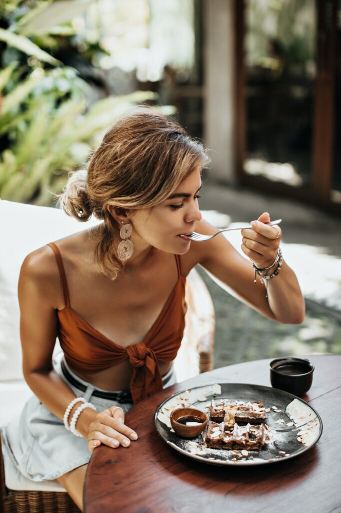 Blonde charming woman in brown bra and denim shorts eats waffle with cream and chocolate sauce and